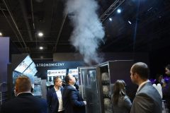 Kilka osób obserwuje przemysłową zmywarkę wypuszczającą parę w kuchni komercyjnej w Expo XXI, otoczoną znakami, sprzętem i marką Hendi podczas Forum Szefów Kuchni i Restauratorów. projekt i nadzór Tatjana Kobuszewska trksystem.com