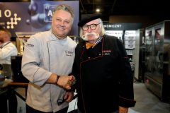 Dwóch szefów kuchni w mundurach podaje sobie ręce i uśmiecha się podczas imprezy kulinarnej, stojąc przed sprzętem kuchennym Hendi i ekspozycjami promocyjnymi w Expo XXI podczas Forum Szefów Kuchni i restauratorów. projekt i nadzór Tatjana Kobuszewska trksystem.com