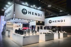 Dobrze oświetlone stoisko targowe prezentuje sprzęt kuchenny i przybory oznaczone marką HENDI Tools for Chefs na wielu dużych banerach. Eleganckie lady prezentują produkty takie jak czajniki, tace i dozowniki. Nowoczesne półki i oświetlenie punktowe tworzą czysty, profesjonalny wygląd. projekt i nadzór Tatjana Kobuszewska trksystem.com