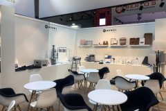 Nowoczesny showroom z białymi ścianami i drewnianymi podłogami prezentuje sprzęt kuchenny Hendi na półkach i ladach. Okrągłe białe stoły otoczone są czarno-białymi krzesłami. Przestrzeń jest dobrze oświetlona, a w tle widoczne są znaki marki gastronomicznej. projekt i nadzór Tatjana Kobuszewska trksystem.com