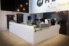 Nowoczesne stoisko wystawowe HENDI z białymi ladami prezentującymi sprzęt kuchenny, świeże owoce i kawę. Nad stoiskiem wiszą geometryczne lampy wiszące. W tle na ścianach znajduje się duży branding i zdjęcia produktów. projekt i nadzór Tatjana Kobuszewska trksystem.com