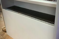 Biała półka ekspozycyjna z broszurami przedstawiającymi szefów kuchni i sprzęt kuchenny w górnym rzędzie oraz starannie ułożonymi czarnymi folderami lub broszurami na dolnej półce. Półka stoi na drewnianej podłodze przy gładkiej ścianie. projekt i nadzór Tatjana Kobuszewska trksystem.com