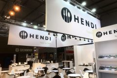 Nowoczesne stoisko wystawowe z dużymi banerami HENDI z logo widelca i łyżki, białymi stołami i krzesłami, broszurami na półkach i jasnym oświetleniem nad głową. W tle widoczne stoiska Tecnoinox i innych marek. projekt i nadzór Tatjana Kobuszewska trksystem.com