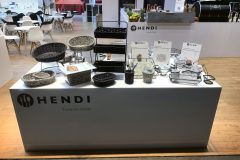 Stół ekspozycyjny z logo Hendi i hasłem Narzędzia dla szefów kuchni, prezentujący różne plecione kosze i metalowe stojaki, znaki informacyjne, przybory i drobne akcesoria kuchenne. W tle widoczne są nowoczesne krzesła i stoły. projekt i nadzór Tatjana Kobuszewska trksystem.com