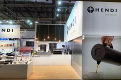 Nowoczesne stoisko wystawowe Hendi, oznaczone jako "Narzędzia dla szefów kuchni". Na stoisku znajdują się eleganckie urządzenia kuchenne, wiszące lampy i ekspozytory produktów. Na ścianach widoczny jest duży branding i zdjęcie czajnika nalewającego kawę. W tle widoczne są inne stoiska. projekt i nadzór Tatjana Kobuszewska trksystem.com