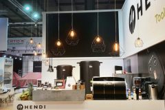 Nowoczesne stoisko wystawowe Hendi obejmuje elegancką białą ladę z ekspresami do kawy i materiałami eksploatacyjnymi. Geometryczne lampy wiszące wiszą powyżej, a duża grafika ścienna prezentuje sprzęt kuchenny. Przestrzeń ma jasną, otwartą atmosferę z drewnianą podłogą. projekt i nadzór Tatjana Kobuszewska trksystem.com