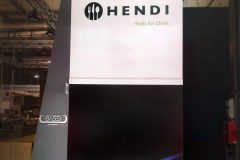 Wysoki wyświetlacz przedstawia logo Hendi z przyborami kuchennymi i hasłem "Narzędzia dla szefów kuchni" na białym tle. Poniżej, obraz palnika kuchennego z niebieskim płomieniem jest pokazany na czarnym panelu wewnątrz nowoczesnej hali wystawowej. projekt i nadzór Tatjana Kobuszewska trksystem.com