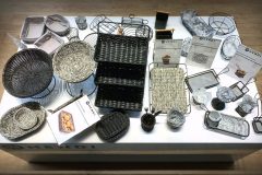 Stół ekspozycyjny z różnymi wiklinowymi i drucianymi koszami, tacami i pojemnikami do serwowania w różnych kształtach i rozmiarach, niektóre z opakowaniami produktów Hendi. Tło stanowi drewniana podłoga, a aranżacja jest schludna i uporządkowana. projekt i nadzór Tatjana Kobuszewska trksystem.com