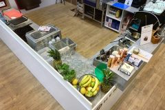 Nowoczesna lada kuchenna w kształcie litery L z ananasami, bananami, cytrynami, limonkami, szklankami do picia, miedzianymi kubkami, przyborami i urządzeniami. Tło obejmuje półki z kubkami, słoikami i różnymi przedmiotami kuchennymi, a wszystko to ustawione na jasnej drewnianej podłodze. projekt i nadzór Tatjana Kobuszewska trksystem.com