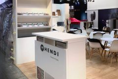 Nowoczesne stoisko wystawiennicze Hendi, prezentujące "Narzędzia dla szefów kuchni". Na stoisku znajdują się białe meble, półki z kolorowymi produktami, lada z broszurami pod spodem oraz miejsca siedzące z białymi krzesłami i drewnianymi stołami na drewnianej podłodze. projekt i nadzór Tatjana Kobuszewska trksystem.com