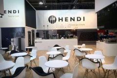 Nowoczesne stoisko wystawowe Hendi z białymi ścianami z dużym logo Hendi, okrągłymi białymi stołami i czarno-białymi krzesłami. Z sufitu zwisają lampy wiszące, a w tle wyświetlane są urządzenia kuchenne. Przestrzeń wygląda schludnie i zachęcająco. projekt i nadzór Tatjana Kobuszewska trksystem.com