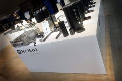 Biała lada ekspozycyjna marki Hendi prezentuje różne urządzenia kuchenne, w tym pojemniki na kawę, dozowniki i grill. Lada jest ustawiona na drewnianej podłodze, a w tle znajdują się znaki informacyjne i inne produkty. projekt i nadzór Tatjana Kobuszewska trksystem.com