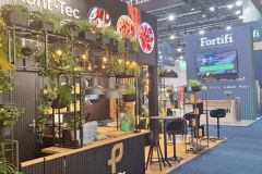 Hala targowa z nowoczesnymi, ozdobionymi roślinami stoiskami, w tym wyróżniającym się czarnym stoiskiem z wiszącą zielenią i górnym oświetleniem. Inne stoiska z brandingiem, miejscami do siedzenia i ekranami cyfrowymi są widoczne wzdłuż niebieskiego chodnika wyłożonego dywanem. projekt i nadzór Tatjana Kobuszewska trksystem.com