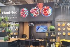 Nowoczesne stoisko wystawowe Plant-Tec Europe zawiera zielone rośliny, czarne i drewniane meble, półki ścienne z butelkami, trzy duże zdjęcia żywności, ekran i okrągłe dekoracyjne talerze na ciemnych ścianach. projekt i nadzór Tatjana Kobuszewska trksystem.com