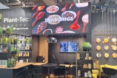 Nowoczesne stoisko targowe dla Plant-Tec Europe firmy LipCo Foods, z zielenią, półkami ze słoikami i butelkami, okrągłymi żółtymi panelami ściennymi, czarnymi lampami wiszącymi i dużym ekranem wyświetlającym przyprawy i żywność. projekt i nadzór Tatjana Kobuszewska trksystem.com