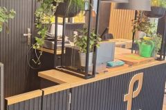 Nowoczesna czarna recepcja z drewnianym blatem i logo "Plant-Tec", otoczona zielonymi roślinami i wiszącymi lampami, tworząca stylową, inspirowaną naturą atmosferę. projekt i nadzór Tatjana Kobuszewska trksystem.com