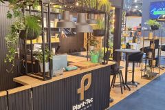 Nowoczesne stoisko wystawowe wyposażone jest w drewnianą i czarną ladę z roślinami doniczkowymi, górnym oświetleniem i stołkami. Widoczne jest logo Plant-Tec, a w tle znajduje się niebieski dywan z innymi stoiskami i ekranem cyfrowym. projekt i nadzór Tatjana Kobuszewska trksystem.com