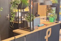 Nowoczesna czarna kabina z drewnianym blatem, ozdobiona wiszącymi i doniczkowymi zielonymi roślinami, lampkami sznurkowymi i logo Plant-Tec z przodu. Na blacie stoi zielona konewka. projekt i nadzór Tatjana Kobuszewska trksystem.com