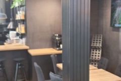 Nowoczesne wnętrze kawiarni z ciemnymi ścianami, drewnianymi podłogami, czarnymi krzesłami, jasnymi drewnianymi stołami, ladą do kawy, półkami z roślinami i żółtą otomaną na pierwszym planie. Pośrodku znajduje się ciemna pionowa kolumna. projekt i nadzór Tatjana Kobuszewska trksystem.com