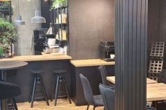 Nowoczesna kawiarnia z czarnymi krzesłami, okrągłymi stolikami, drewnianą podłogą i czarną ladą. Przestrzeń zdobią rośliny i lampy wiszące. Powyżej widnieje napis Plant-Tech Europe z czerwono-białym logo. projekt i nadzór Tatjana Kobuszewska trksystem.com