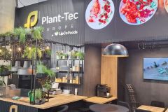 Nowoczesne stoisko wystawiennicze dla Plant-Tec Europe firmy LipCo Foods obejmuje zieleń, półki z roślinami, małą ladę, czarne krzesła i stoły, górne oświetlenie oraz ścianę wyświetlającą kolorowe zdjęcia żywności i duży ekran. projekt i nadzór Tatjana Kobuszewska trksystem.com