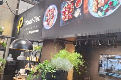 Stoisko targowe dla Plant-Tec Europe z zielenią, sprzętem do gotowania i dużymi wyświetlaczami pokazującymi platerowane potrawy; nowoczesny, elegancki design z czarnymi i drewnianymi akcentami. projekt i nadzór Tatjana Kobuszewska trksystem.com