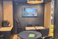 Nowoczesne wnętrze kawiarni z drewniano-czarnym wystrojem, małymi stolikami i krzesłami, ekspresem do kawy na ladzie, żółtą sofą i ekranem telewizora na ścianie wyświetlającym kobietę posługującą się językiem migowym. projekt i nadzór Tatjana Kobuszewska trksystem.com