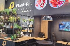 Nowoczesne stoisko w stylu kawiarni z oznakowaniem Plant-Tec Europe, bujnymi zielonymi roślinami, półkami z oświetleniem i ladą. Na ekranie telewizora wyświetlany jest industrialny obraz, a powyżej znajduje się duże zdjęcie jedzenia. Czarne krzesła i stoły są starannie ustawione. projekt i nadzór Tatjana Kobuszewska trksystem.com