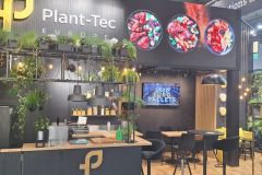 Nowoczesne stoisko wystawiennicze Plant-Tec Europe z bujnymi zielonymi roślinami, drewnianymi półkami, czarnymi meblami, ladą i wyświetlaczem ściennym z kolorowymi obrazami żywności i ekranem telewizyjnym. projekt i nadzór Tatjana Kobuszewska trksystem.com