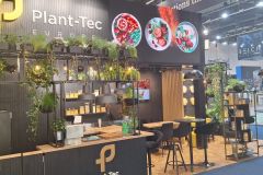 Stoisko targowe dla Plant-Tec Europe, udekorowane zielenią i roślinami, drewnianymi akcentami, wysokimi krzesłami i stołami. Na tylnej ścianie wyświetlane są okrągłe obrazy potraw. Stoisko jest dobrze oświetlone i zachęcające. projekt i nadzór Tatjana Kobuszewska trksystem.com