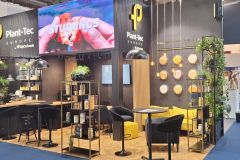 Nowoczesne stoisko wystawowe Plant-Tec Europe zawiera czarno-żółte meble, zielone rośliny, oświetlenie ścienne i duży ekran wyświetlający obraz rąk trzymających jedzenie z napisem STUFFINGS. projekt i nadzór Tatjana Kobuszewska trksystem.com