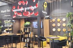Nowoczesne stoisko wystawowe firmy Plant-Tec Europe charakteryzuje się eleganckim wystrojem w czerni i drewnie, zielonymi roślinami, cyfrowym ekranem wyświetlającym SYSTEMY PIECZENIA oraz miejscami do siedzenia z żółtymi akcentami na targach. projekt i nadzór Tatjana Kobuszewska trksystem.com