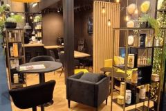 Nowoczesne stoisko wystawiennicze dla Plant-Tec Europe obejmuje eleganckie czarne i drewniane meble, półkę ekspozycyjną z produktami, wiszące oświetlenie oraz przytulną część wypoczynkową z krzesłami i małym stolikiem. projekt i nadzór Tatjana Kobuszewska trksystem.com