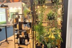 Stoisko targowe prezentuje zielone rośliny doniczkowe i wiszące pnącza na półkach, z pudełkami produktów ułożonymi obok. Stoisko ma czarne ramy i drewniane panele i jest otoczone innymi ekspozycjami wystawowymi. projekt i nadzór Tatjana Kobuszewska trksystem.com