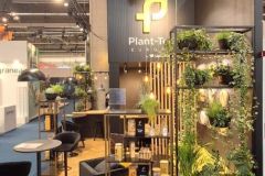 Stoisko na targach z marką Plant-T, z półkami z roślinami doniczkowymi, słoikami i zielonymi dekoracjami. Pod industrialnym sufitem znajdują się wysokie stoły, taborety i wysokie oświetlenie. projekt i nadzór Tatjana Kobuszewska trksystem.com