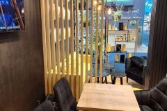 Nowoczesne wnętrze kawiarni z drewnianymi stołami, czarnymi wyściełanymi krzesłami, przegrodą z pionowymi drewnianymi listwami i dekoracyjnymi półkami. Jaskrawożółte siedzenia i znak "Fortifi" są widoczne przez przegrodę. projekt i nadzór Tatjana Kobuszewska trksystem.com