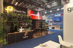 Stoisko targowe Plant-Tec Europe z zielenią, drewnianymi półkami z roślinami i słoikami, ladą ze stołkami i ekranami cyfrowymi. W pobliżu hali wystawowej znajdują się również stoiska firm Fortifi i Grant. projekt i nadzór Tatjana Kobuszewska trksystem.com
