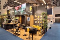 Nowoczesne stoisko wystawowe z drewnianą podłogą, czarno-żółtym wystrojem, zielonymi roślinami, okrągłymi wiszącymi lampami i brandingiem Plant Pro. Wokół znajdują się szerokie, wyłożone niebieską wykładziną chodniki i inne stoiska wystawowe. projekt i nadzór Tatjana Kobuszewska trksystem.com