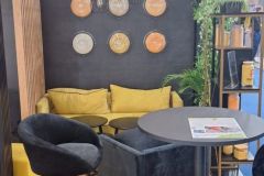 Nowoczesne stoisko targowe zawiera żółto-czarną kanapę, czarny fotel, okrągły czarny stół z czasopismem, czarny stołek barowy, dekoracyjne zegary ścienne, wiszące lampy, rośliny i drewnianą podłogę. projekt i nadzór Tatjana Kobuszewska trksystem.com
