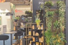 Stoisko targowe o nowoczesnym wyglądzie, z czarnymi meblami, zielonymi roślinami doniczkowymi, półkami z pudełkami i słoikami oraz żółtym logo na ciemnej ścianie w tle. projekt i nadzór Tatjana Kobuszewska trksystem.com