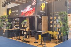 Nowoczesne stoisko wystawiennicze Plant-Tec Europe obejmuje zieleń, drewniane półki z produktami, bar ze stołkami i duży ekran cyfrowy wyświetlający słowo MARINADES. Wokół znajdują się inne stoiska. projekt i nadzór Tatjana Kobuszewska trksystem.com