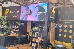 Nowoczesne stoisko wystawiennicze Plant-Tec Europe charakteryzuje się czarno-żółtą kolorystyką, wiszącymi roślinami, wyświetlaczami produktów, zegarami ściennymi i dużym ekranem cyfrowym wyświetlającym PRODUKTY NUTRITION AND SUPPLEMENTS. projekt i nadzór Tatjana Kobuszewska trksystem.com