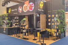 Nowoczesne stoisko targowe dla Plant-Tec Europe charakteryzuje się czarno-żółtym wystrojem, zielonymi roślinami, półkami ekspozycyjnymi, stołkami barowymi i podświetlanymi okrągłymi obrazami roślin i produktów nad ladą. projekt i nadzór Tatjana Kobuszewska trksystem.com