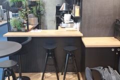 Nowoczesna kawiarnia z czarnymi stołkami przy drewnianej ladzie, roślinami na półkach, lampami wiszącymi i czarnym krzesłem z plecakiem; jasna drewniana podłoga w jodełkę i betonowy filar w tle. projekt i nadzór Tatjana Kobuszewska trksystem.com