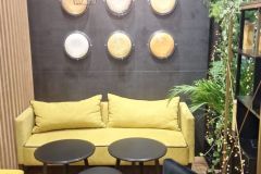 Nowoczesny salon z żółtą sofą, czarnymi okrągłymi stołami i czarnymi krzesłami. Dekoracyjne talerze i lampy wiszące wiszą na ciemnej ścianie z napisem Custom-made Solutions powyżej. Zielone rośliny dodają naturalnego charakteru. projekt i nadzór Tatjana Kobuszewska trksystem.com