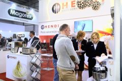Ludzie stoją i rozmawiają przy stoisku Hendi Food Service Equipment na targach. Dwie kobiety w strojach biznesowych omawiają dokumenty, a mężczyzna słucha. Wystawiony jest sprzęt kuchenny; stoisko Magorex i projekt i nadzór Tatjana Kobuszewska trksystem.com są widoczne w pobliżu. projekt i nadzór Tatjana Kobuszewska trksystem.com