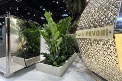 Zbliżenie na metalowy ekspres do kawy La Pavoni z geometrycznym wzorem, obok lustrzanej donicy z zielonymi roślinami liściastymi, wszystko ustawione na białej powierzchni w jasnym oświetleniu wewnętrznym. projekt i nadzór Tatjana Kobuszewska trksystem.com