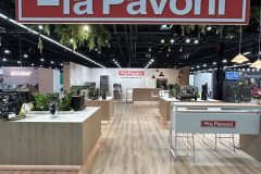 Nowoczesny, dobrze oświetlony showroom z drewnianymi podłogami prezentuje ekspresy do kawy. Duży czerwono-biały znak la Pavoni wisi nad głową, a na ladach w całej przestrzeni znajduje się pasujący branding. Zielone rośliny zdobią lady. projekt i nadzór Tatjana Kobuszewska trksystem.com