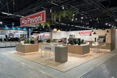 Nowoczesne stoisko wystawowe La Pavoni prezentuje ekspresy do kawy i sprzęt kawowy na drewnianych ladach, z zielenią nad głową i dużym czerwonym znakiem La Pavoni zawieszonym nad stoiskiem. projekt i nadzór Tatjana Kobuszewska trksystem.com