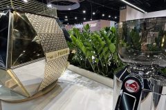 Nowoczesny ekspres do kawy z błyszczącym, złotym, geometrycznym wzorem stoi obok chromowanego młynka do kawy na białej ladzie, z zielonymi roślinami i znakami wystawowymi widocznymi w tle. projekt i nadzór Tatjana Kobuszewska trksystem.com